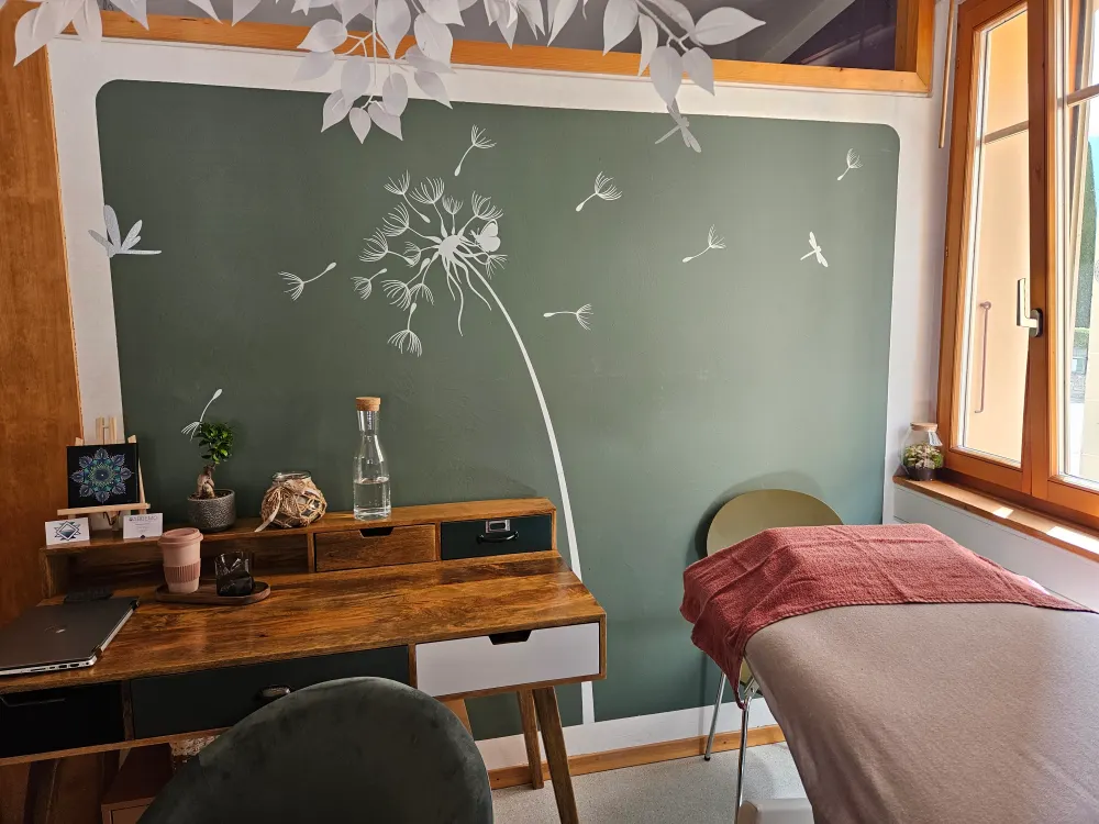 Le cabinet d’Abbemo à Sierre : un bureau en bois et une table de soin devant un mur vert orné d’un grand pissenlit.